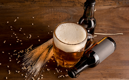 Кружка пива с пеной, две бутылки и колосья пшеницы на деревянном столе